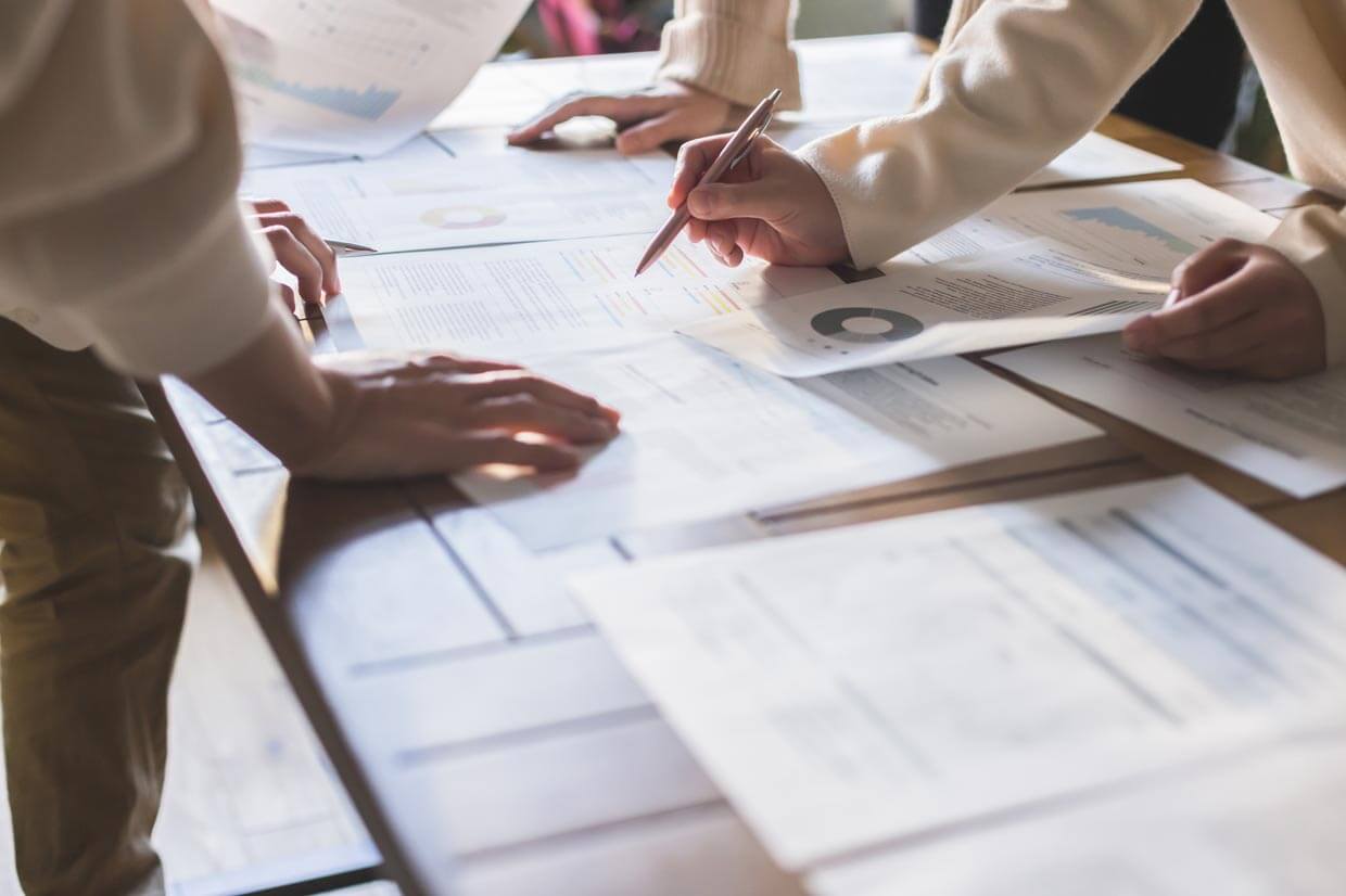 書類を確認する複数の手： 机の上に広げられた多くの資料を、複数人で書き込みながら確認。
